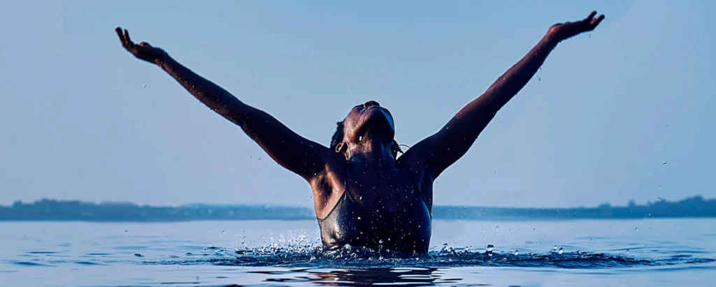 Mujer con los brazos alzados dentro del agua, de espaldas al horizonte