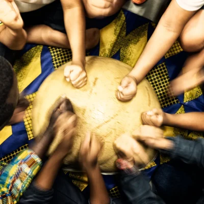 Muchas manos de distintas personas tocando a la vez la piel de un mismo tambor, rodeadas de telas africanas de colores