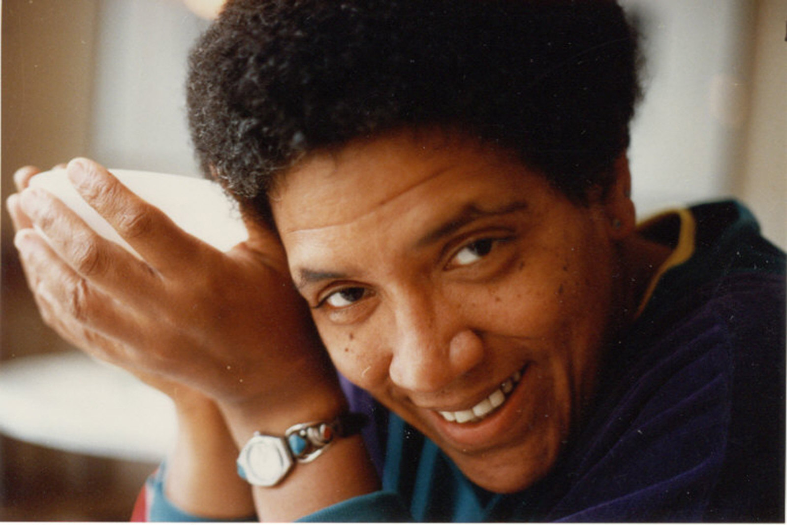 Audre Lorde sonriendo en un plano cercano, años 80, con cabello afro corto y una prenda azul con detalles amarillos, apoyando la cabeza en su mano.