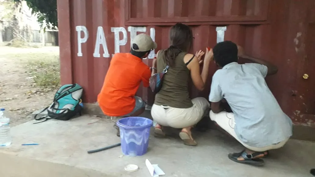 Tres personas agachadas pintando a mano las letras de «Papelaria» en el contenedor