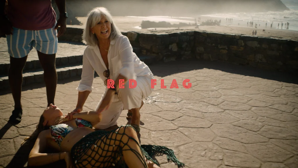 Fotograma de Red Flag, de Manuel Martín Merino, amb el títol sobreimprès: una dona demana ajuda al costat d'una jove estesa en un passeig vora el mar
